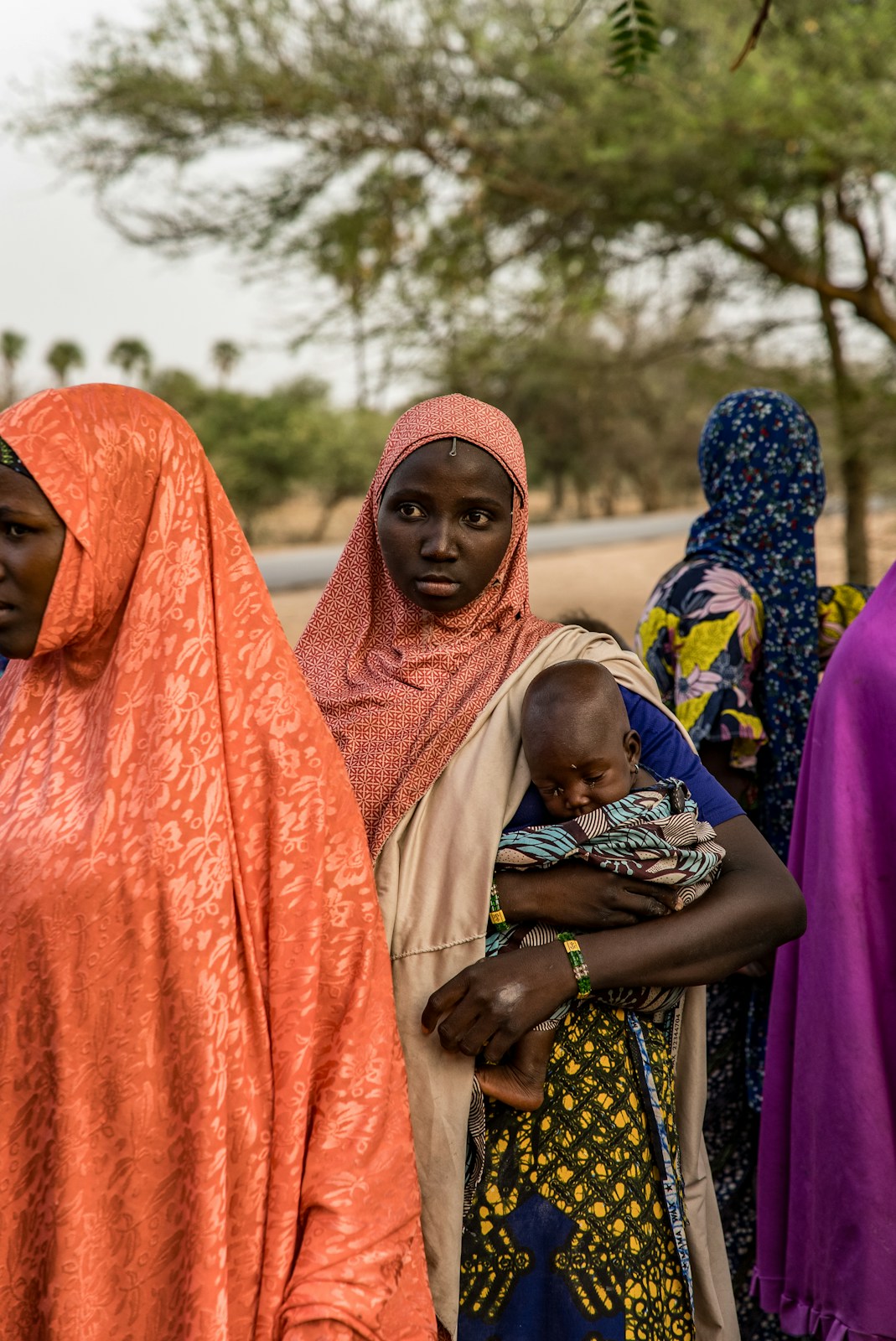 A woman in a pink headscarf holds a baby outdoors