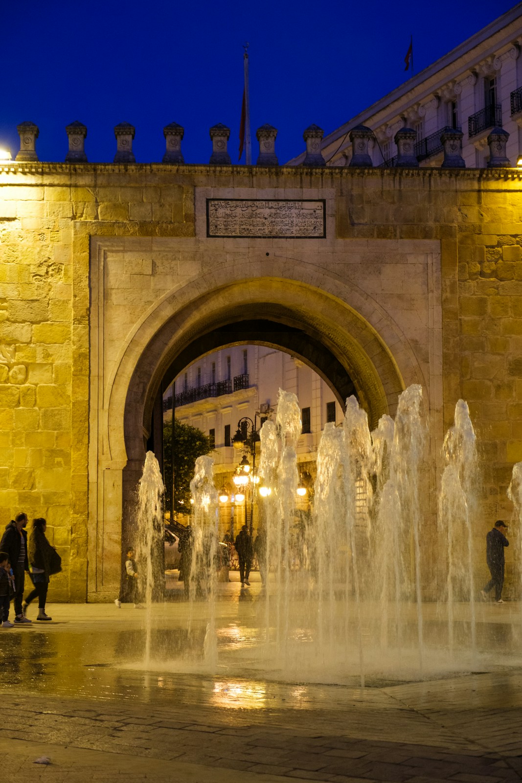 People walk past a fountain near an ancient stone archway